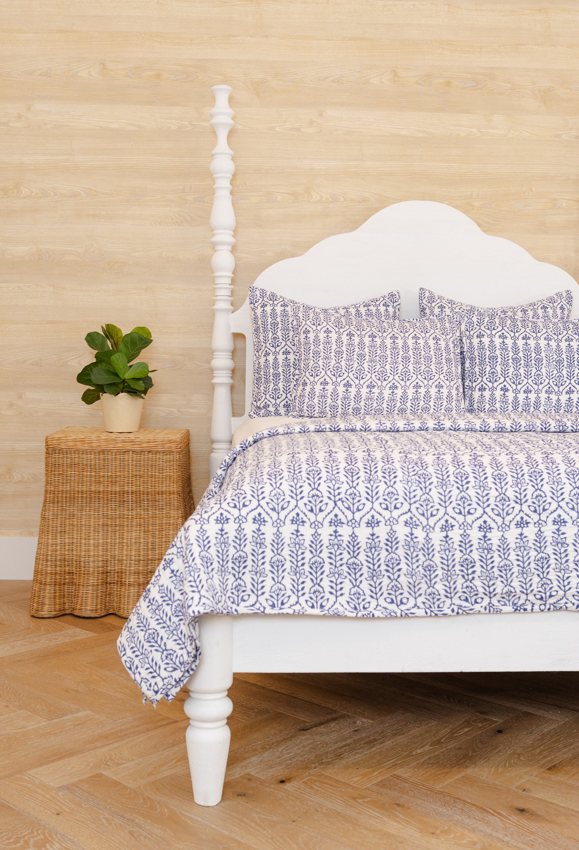 Bed with patterned bedding in a room with wooden floor and wicker side table.